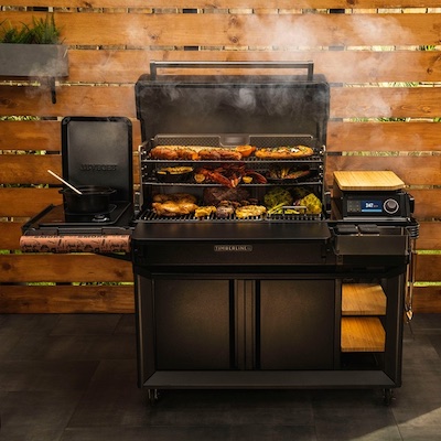 A Traeger grill near a wall with open lid and food cooking.