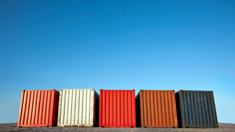 Five colorful shipping containers sit on the ground under a clear blue sky.