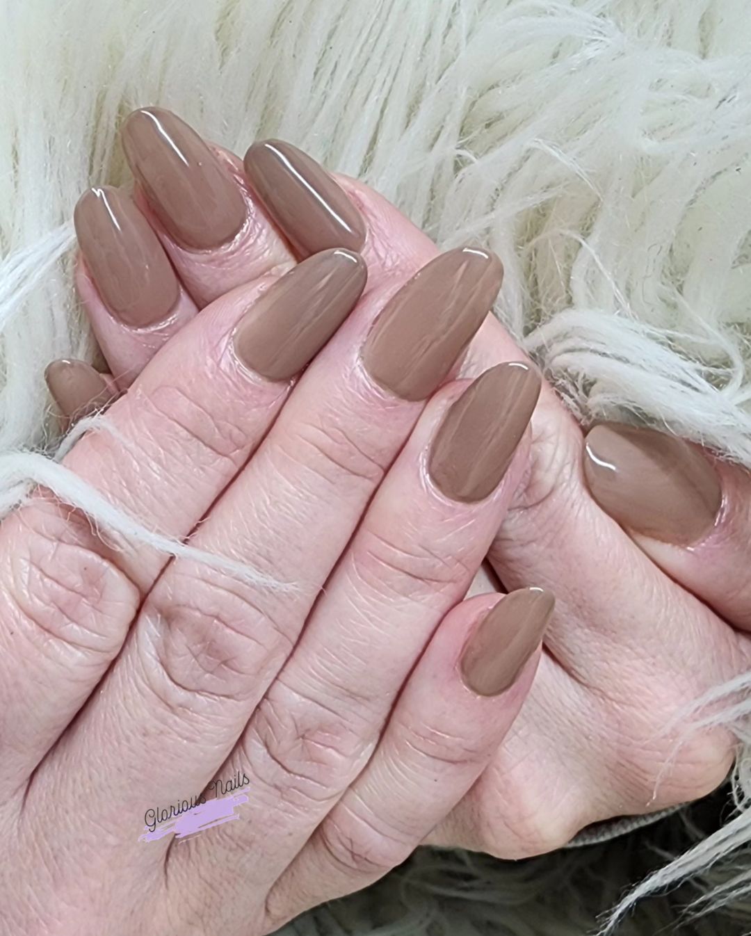 Two hands with long, almond-shaped, taupe-colored nails resting on a fluffy white surface.



