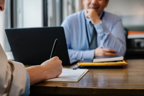 businessman writing on note with pen over the wooden desk with laptop facing the interviewer