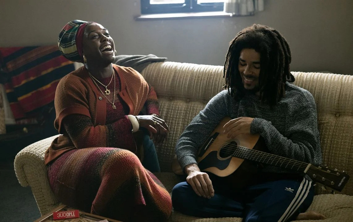 Escena de la película de Bob Marley donde dos personajes, uno tocando la guitarra y otro riendo, comparten un momento en un sofá.