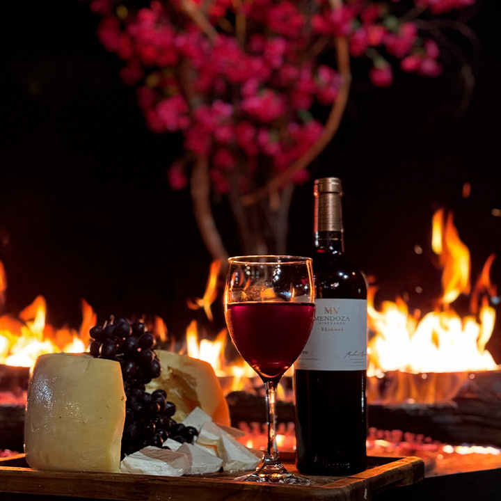 Vinho tinto em taça, queijo, uvas e vinho em uma mesa ao lado de uma fogueira ao ar livre à noite, com árvore de flores rosas ao fundo, criando uma atmosfera aconchegante e romântica para jantar ao ar livre.