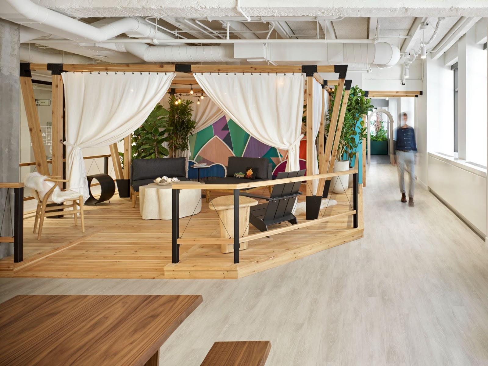 A brightly lit office space features a wooden platform with white tent-like curtains, a colorful mural background and low black chairs and tables for work.