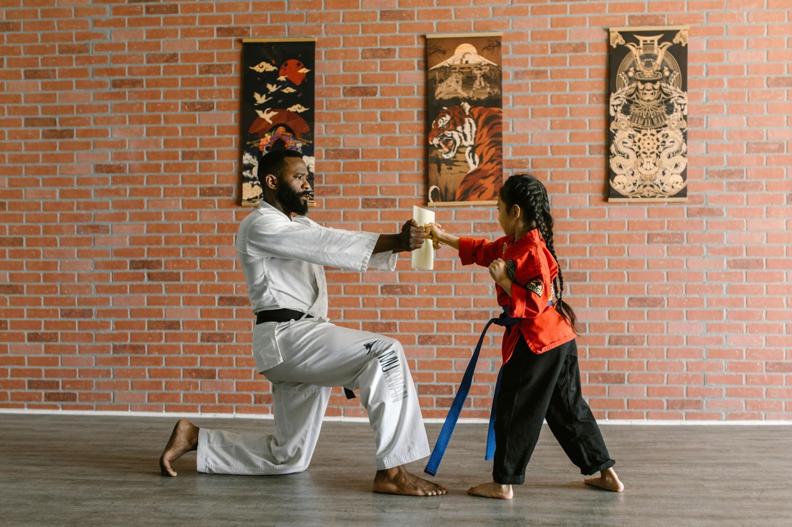 A martial arts instructor kneels on one knee, holding a piece of paper, while a young student stands ready to hit it.