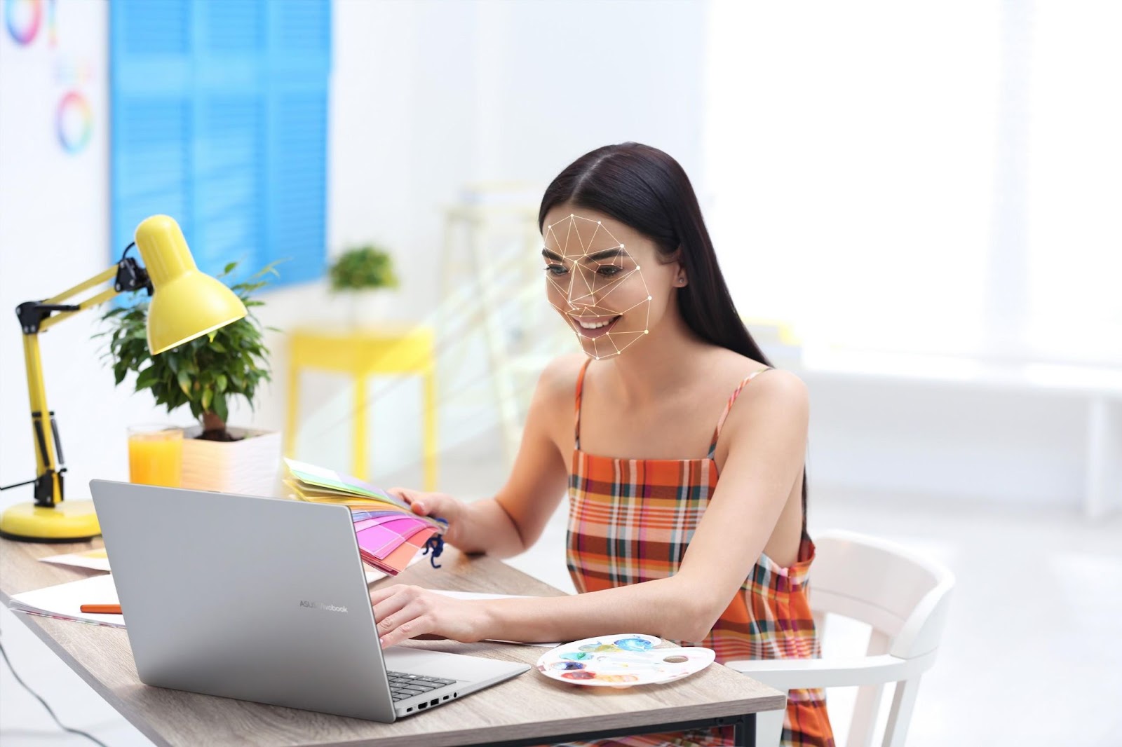 Mulher usando o ASUS Vivobook 16 para trabalho criativo, com paleta de cores e materiais de arte ao lado, em ambiente bem iluminado e colorido.