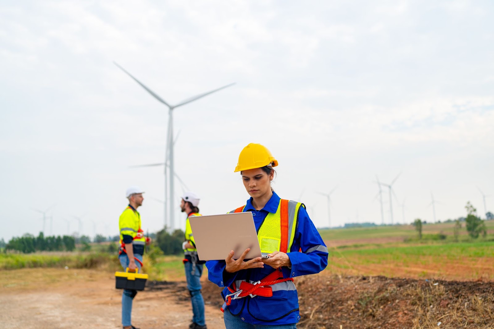 Woman in a hard hat using a laptop next to wind turbines. 