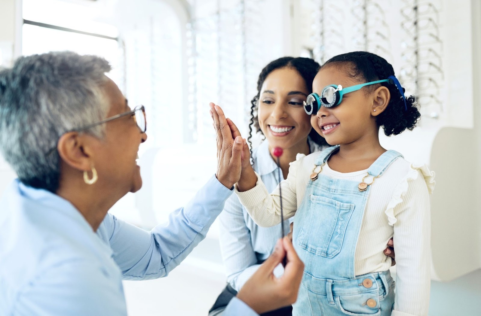 A laughing optometrist high-fiving a young girl during an eye exam while her mother smiles.