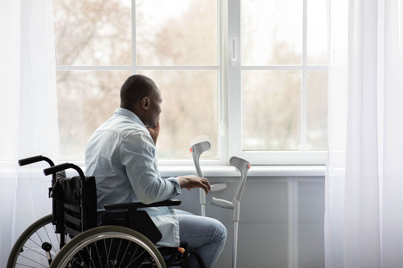 A man sits in a wheelchair, with a pair of crutches next to him, looking out the window after he suffered a hip injury in a car crash.