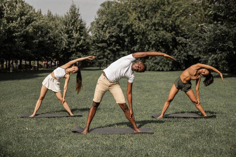 Three individuals are performing a side stretch exercise on yoga mats in an outdoor park setting. They are standing on grass, with trees in the background, and are bending sideways with one arm extended overhead and the other arm reaching down towards their legs.