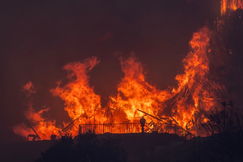 A raging wildfire consumes a structure, with massive flames engulfing the scene. Bright orange and yellow fire contrasts against the dark sky, creating an intense and dramatic visual. A fence and some vegetation are silhouetted in the foreground, emphasizing the destructive power of the blaze.