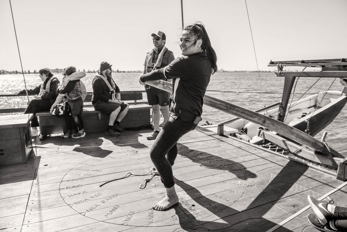 A gray and white photo of a woman navigating the Hinemoana, with five people in the background looking relaxed.