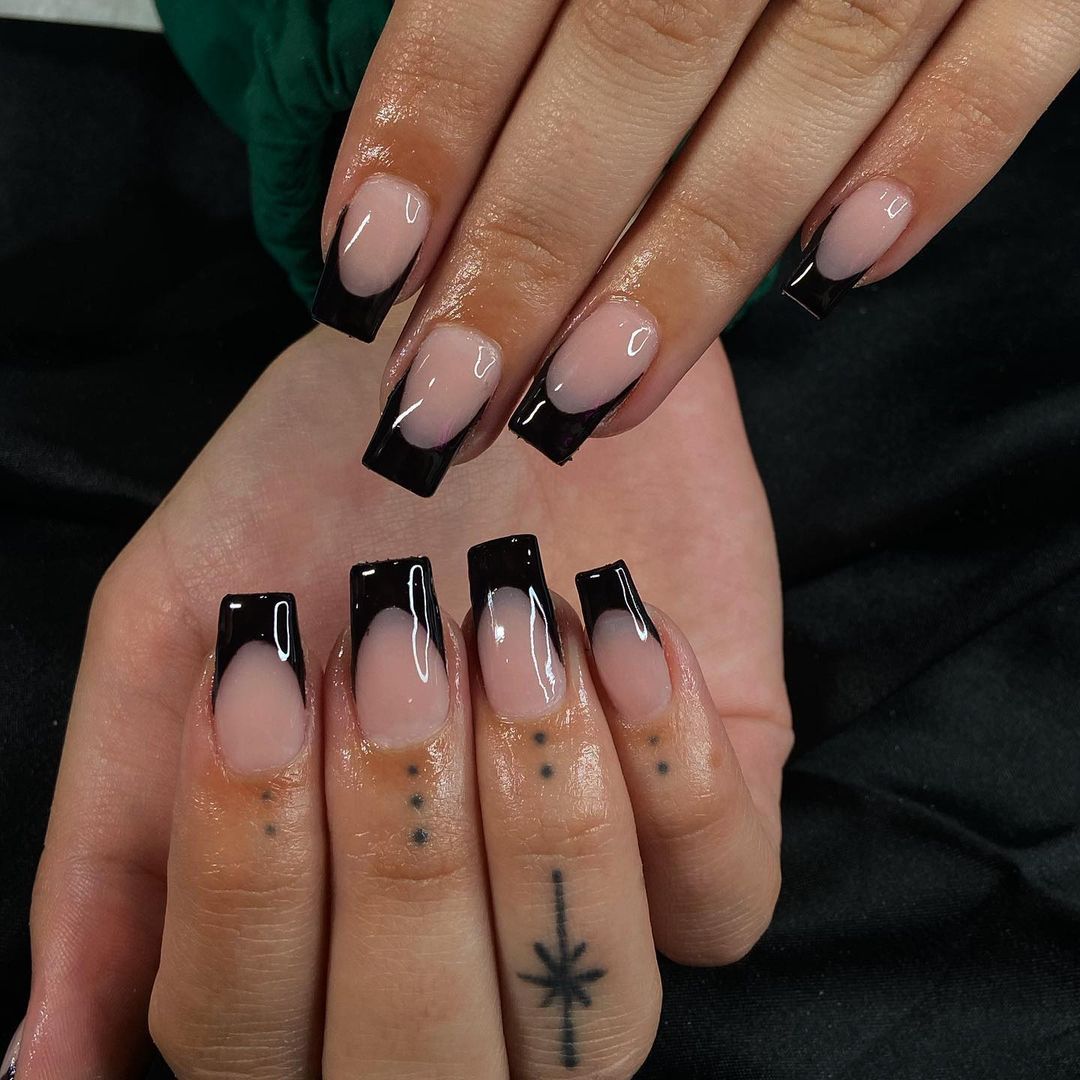 Close-up of a person's hands with nude, glossy, square-shaped nails, black French tips, dot tattoos, and star-like tattoo.