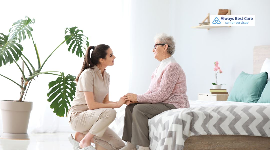 A caregiver kneels beside a senior woman seated on a bed in a softly lit bedroom, offering comfort and compassionate emotional support.
