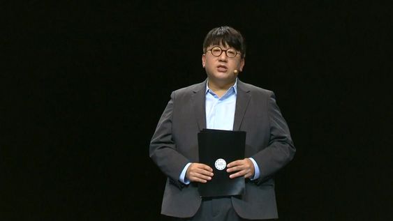 A picture of Bang Si Hyuk wearing a dark ash color suit 