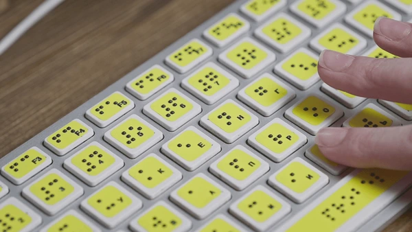 A close-up of a braille keyboard for a computer, placed on a wooden desk beside mobility aids such as white canes.