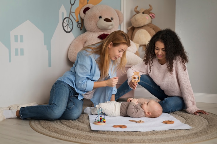 Deux mamans jouant avec leur bébé dans une chambre décorée de peluches et jouets au sol.