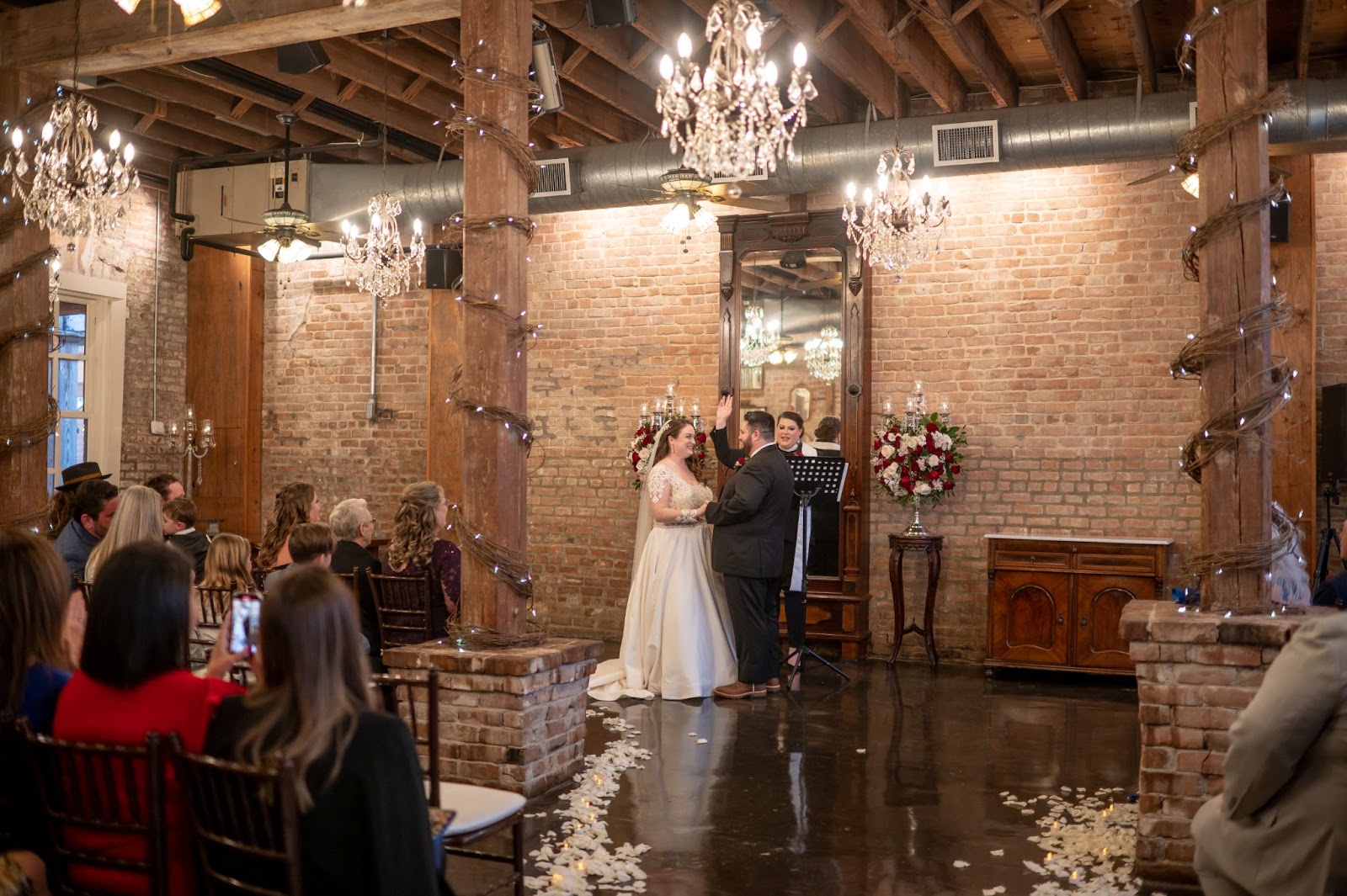 ALT TEXT: In Galveston County's premiere wedding venue, Butler’s Courtyard, a bride and groom exchange heartfelt vows, surrounded by elegant custom floral arrangements. The aisle is lined with white rose petals. Guests sit comfortably in padded Chiavari chairs, sheltered from the elements, enhancing the intimate and elegant atmosphere of the ceremony. The Grand Hall is the best wedding ceremony location between Houston and Galveston.