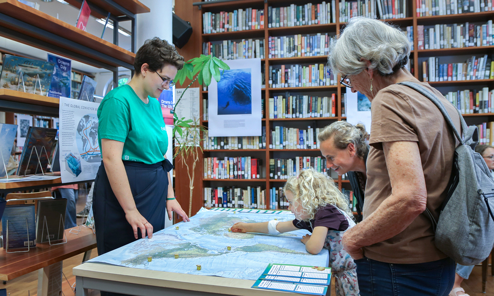 A woman wearing a green T-shirt stands in front of a table with a large world map spread out on top. A little girl is pointing to a spot on the map, while two adults look on. Bookshelves are visible behind the group. 