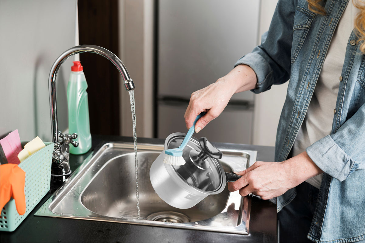 Seorang wanita mencuci panci stainless di wastafel dapur, menjaga kebersihan dengan cara yang higienis dan mudah. 