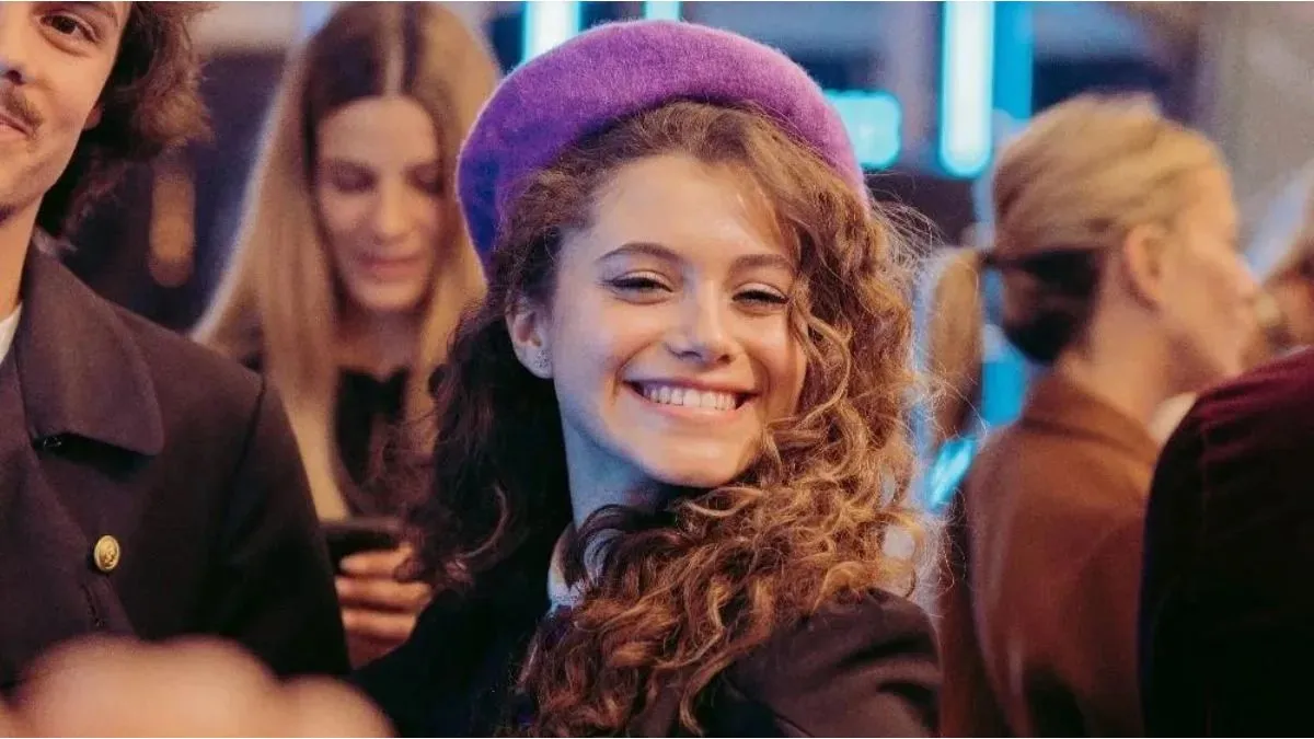 Joven con boina púrpura y cabello rizado sonriendo en un evento social, rodeada de personas.