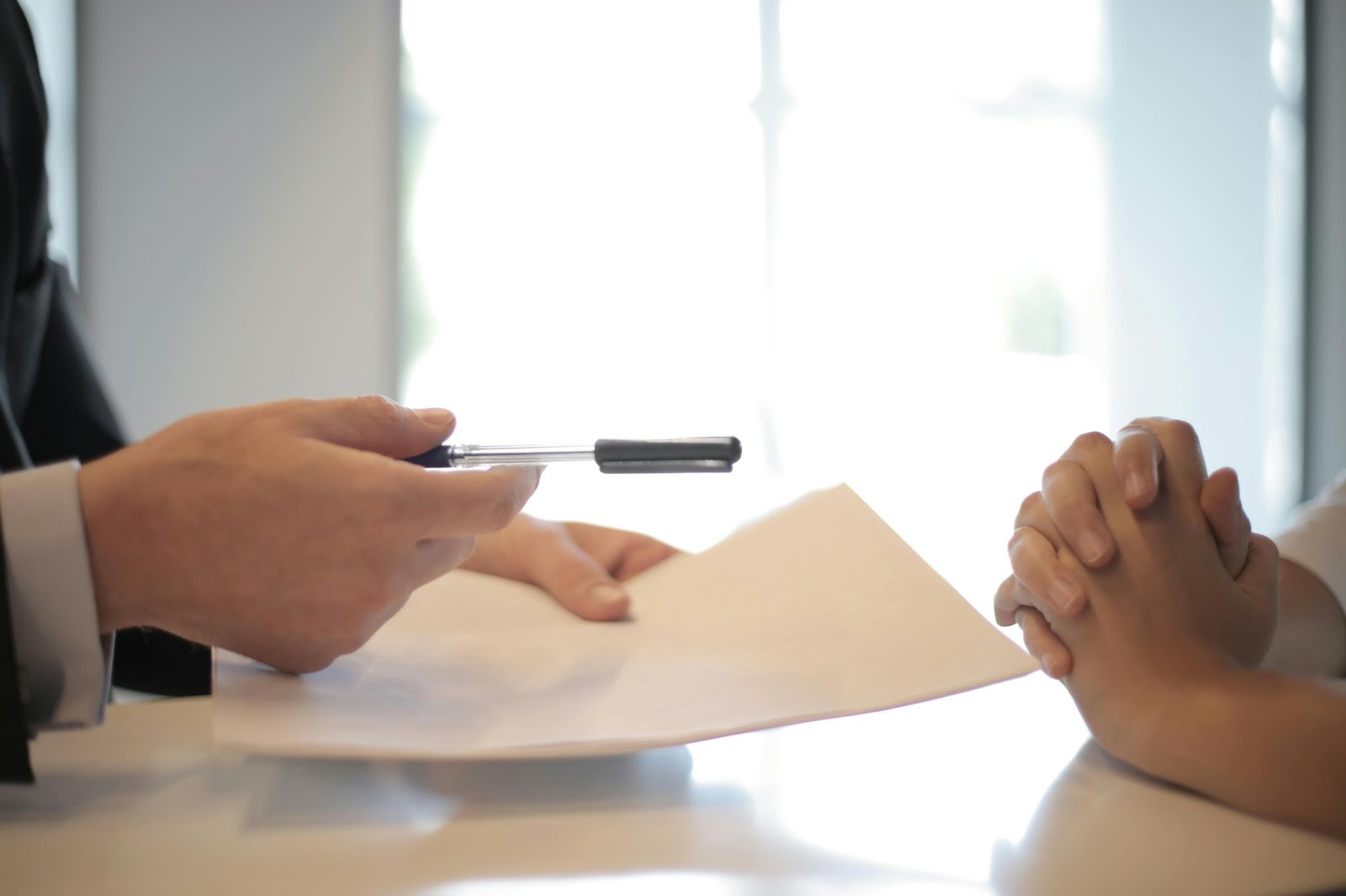 person with hands clasped while another person offers pen and papers to sign