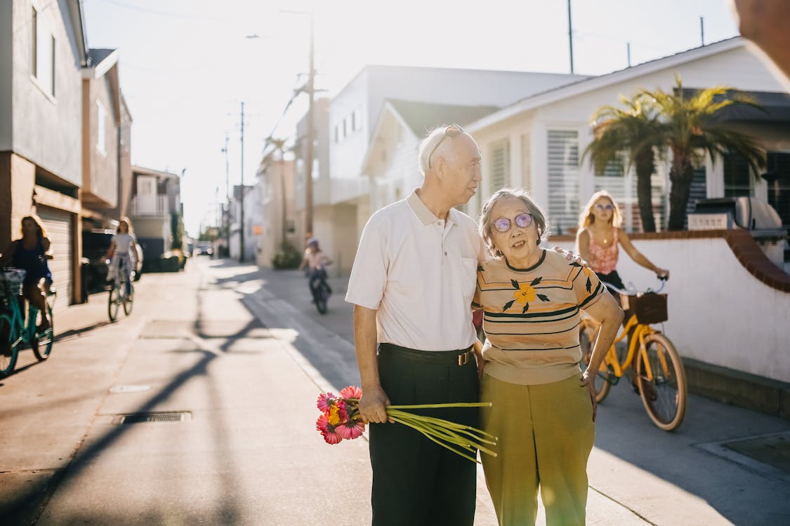 Free Elderly couple enjoying a walk with flowers in a sunny street setting, surrounded by cyclists. Stock Photo