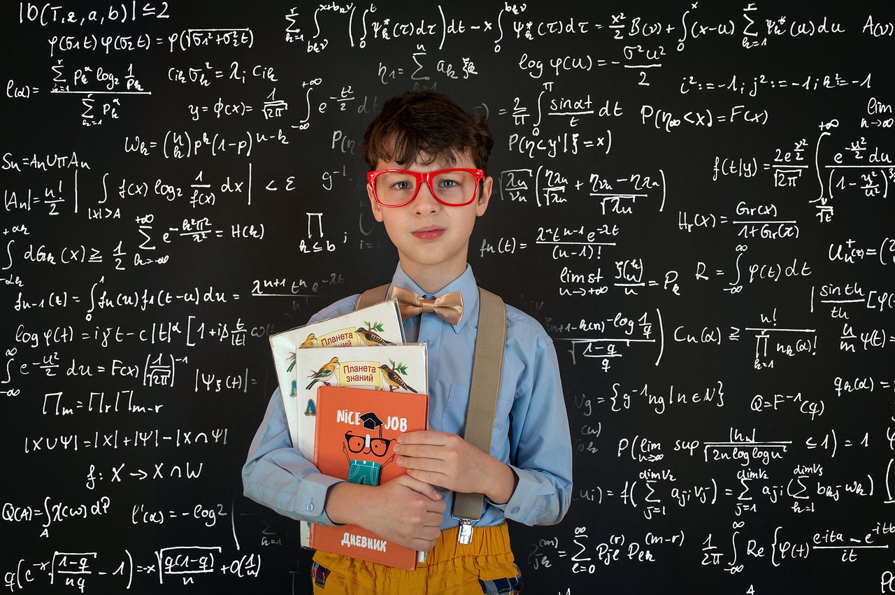 A young boy in glasses stands in front of a blackboard with math equations. 