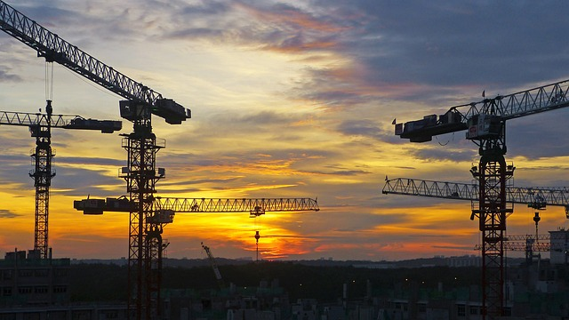 Silhouetted construction cranes against a dramatic sunset sky with orange and blue hues symbolize the progress of sustainable public infrastructure.