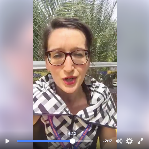 Tarzan wears big nerdy glasses, bright red lipstick and a cool as fuck checker patterned dress with a huge collar. Palm fronds in the background, as she talks to the camera. 