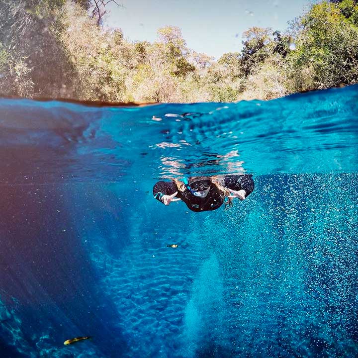Mergulhador em traje de mergulho, nadando na água cristalina de um rio rodeado por árvores verdes e céu azul acima, praticando mergulho esportivo.