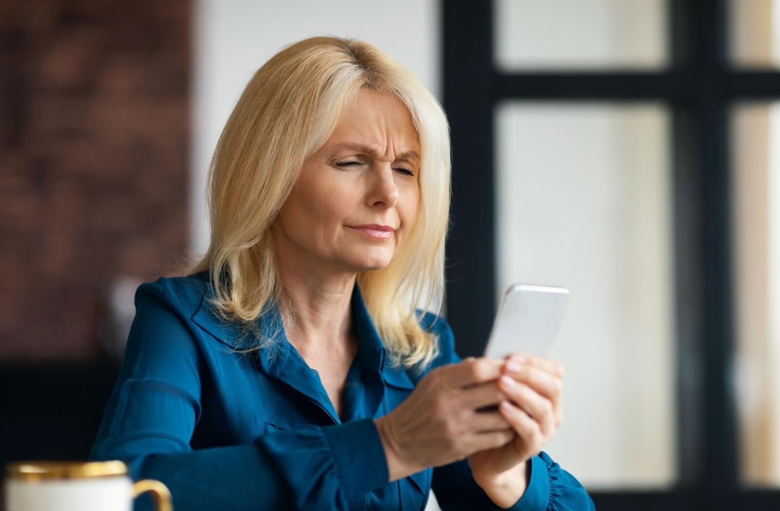 A person squinting while holding their phone at arm’s length, struggling to read small text due to hyperopia (farsightedness).
