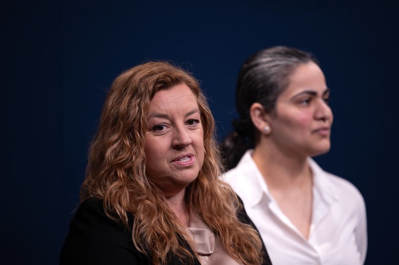 A close-up view of a lawmaker, with long auburn-colored hair and wearing a black blazer with a champagne-colored shirt, as they speak at a press conference. Another lawmaker with salt and pepper hair and wearing a white blouse can be seen standing nearby.