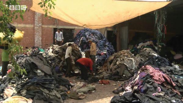 Letter B. Huge piles of unwanted clothes sit on the ground outside a factory. Men are organizing the clothes into piles by the type of clothing.