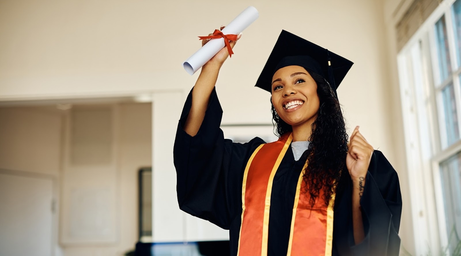 Smiling nutrition graduate in cap and gown holding a diploma and celebrating in a brightly lit room.