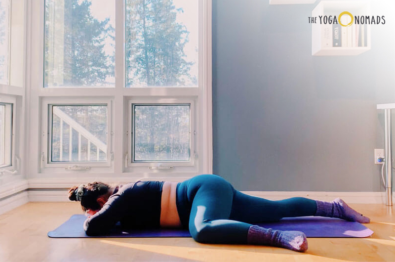 Restorative Half Frog Pose: An individual is performing a yoga pose on a purple mat inside a room with hardwood floors and large windows that reveal trees outside. The individual is wearing blue leggings, a dark top, and purple socks.