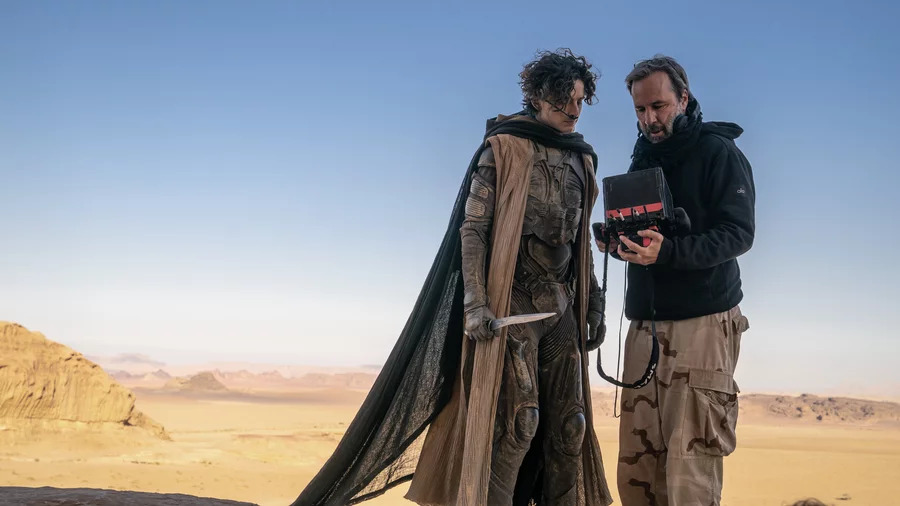 Actor vestido con un traje futurista y director revisando un monitor en el set de rodaje de una película en un paisaje desértico.