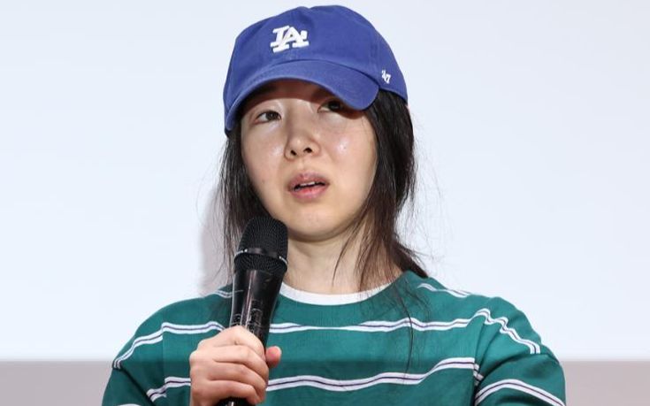 This contain an image of  Min Hee Jin holding a microphone in front of her face and wearing a baseball cap