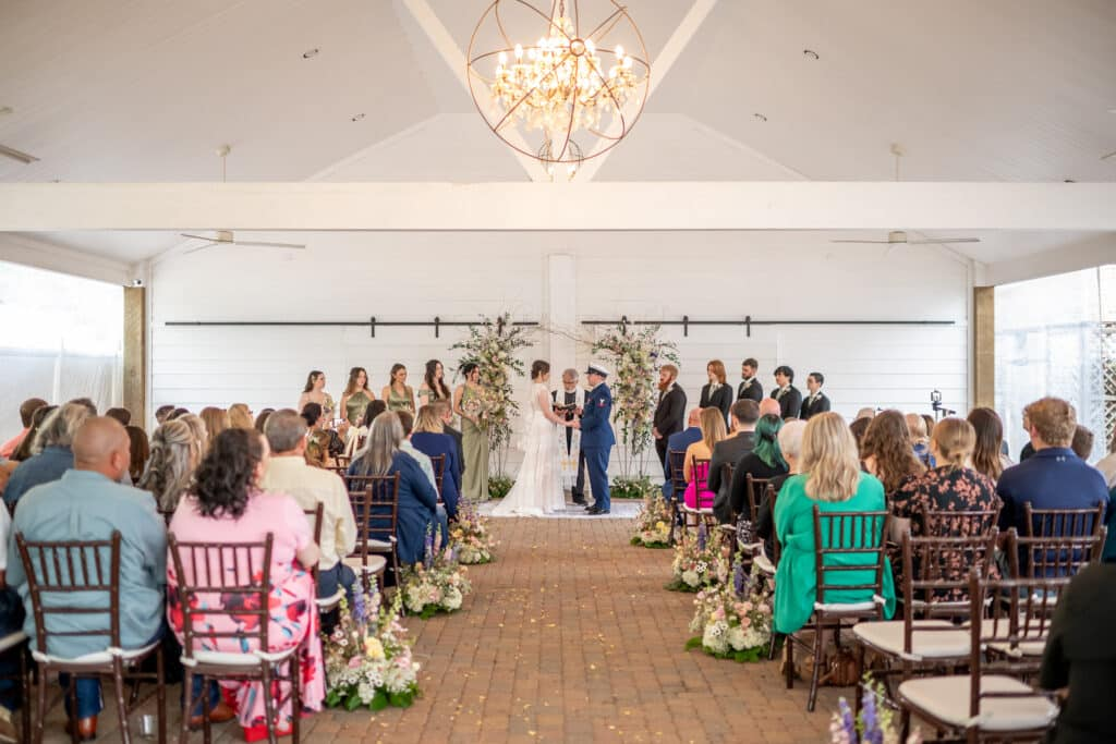 Bride and military groom share their vows in the bright, white, and airy Pavilion, one of many perfect wedding ceremony locations at Butler's Courtyard. The open-air structure offers a perfect blend of shade and outdoor ambiance, highlighting the venue's elegance and unique charm as a premier wedding ceremony location near Houston, Texas.