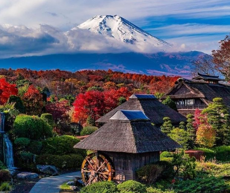 A house with a thatched roof and a water wheel in front of a mountainDescription automatically generated