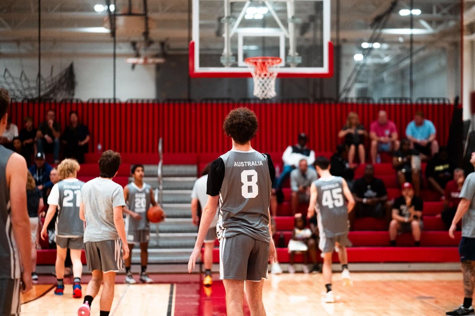 Several basketball players compete on the court with Australia jersey bibs on. None of their faces are visible as they are all looking at the hoop on the far end of the court.