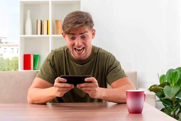 Excited young blonde handsome man sits at table with cup holding and looking at phone inside living room