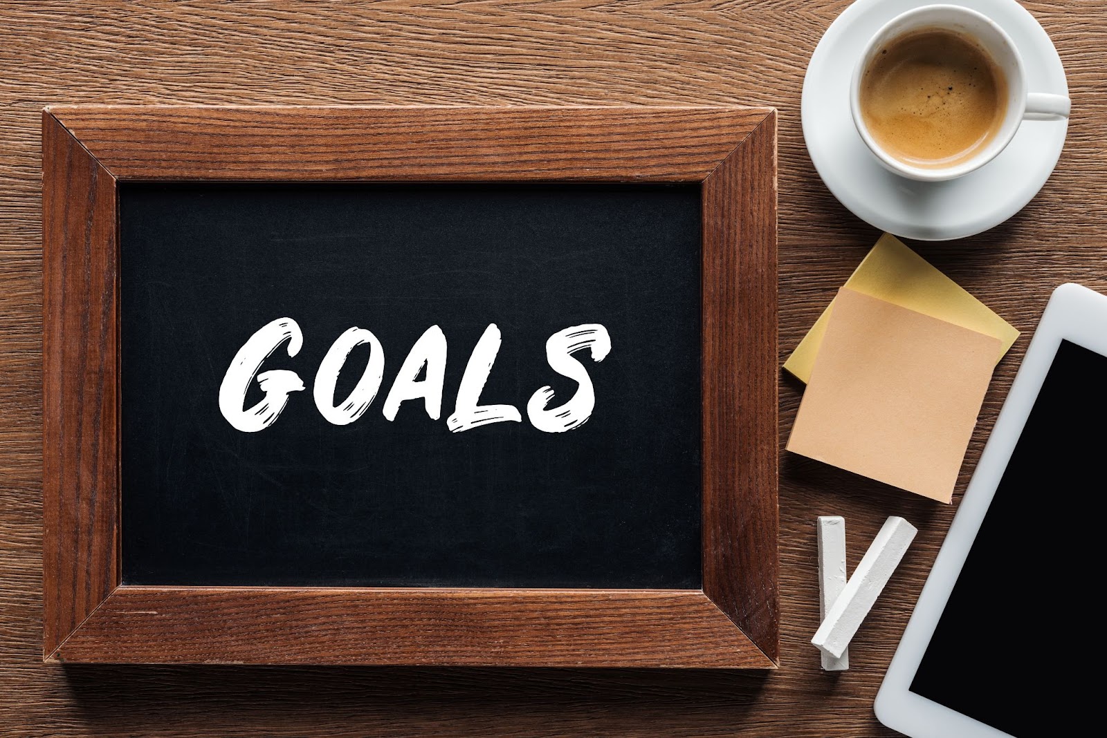  Chalkboard with the word “goals” next to chalk, a tablet, sticky notes, and a cup of coffee.