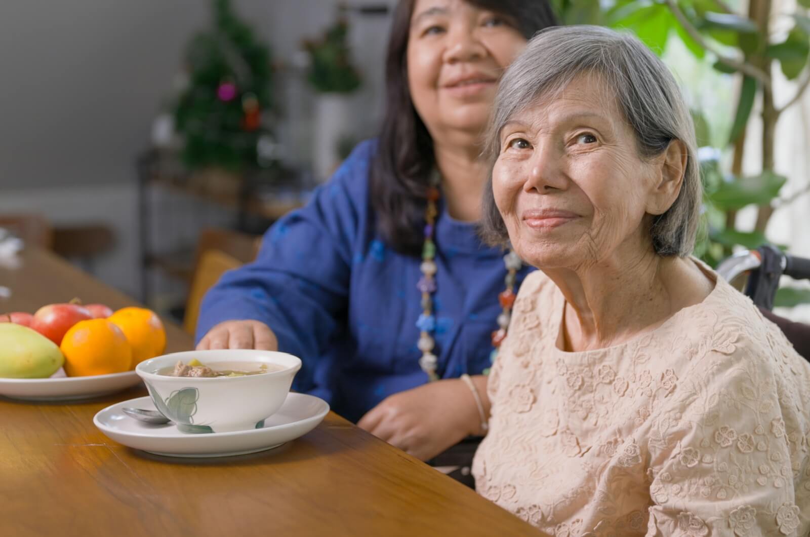 An adult child sitting at a dining table next to a smiling parent with dementia during meal time.
