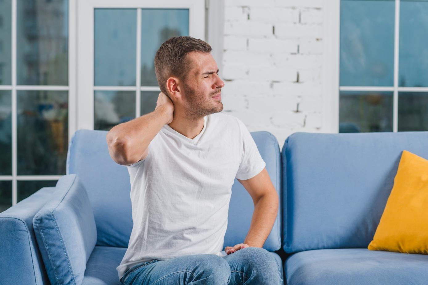 Man sitting on a couch holding his neck and wincing in pain, illustrating symptoms of whiplash