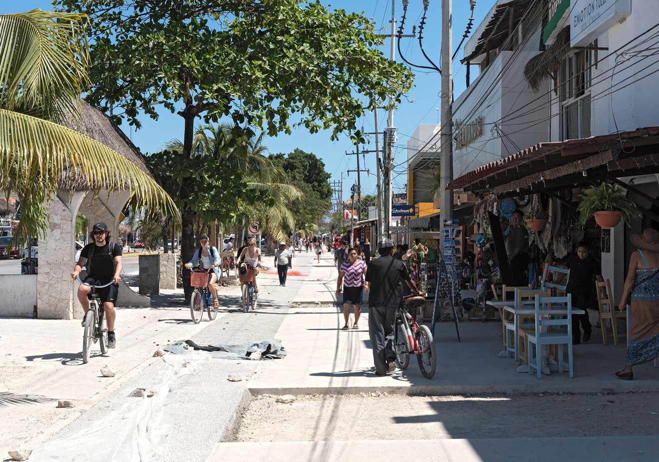 People walking and cycling on a sunlit street lined with shops on one side and trees on the other.