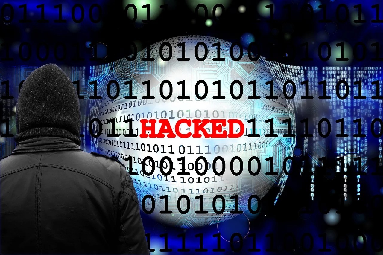 Person on a black hoodie facing towards a background with a “HACKED” written boldly in red and with cryptographic numbers 