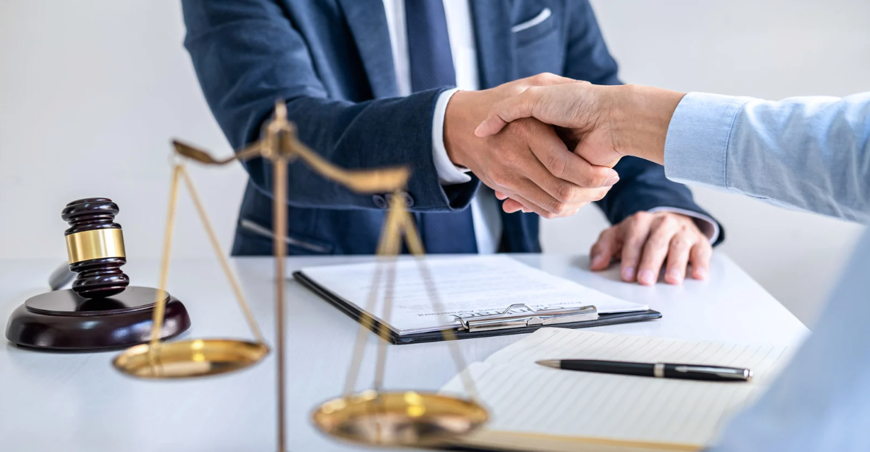 (A lawyer and client shake hands at a desk with legal documents, a gavel, and scales of justice, symbolizing a successful legal agreement or consultation.)