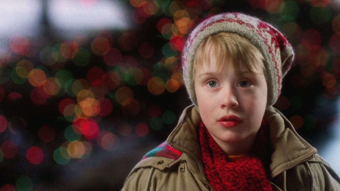 Kevin McCallister con gorro de invierno y bufanda roja, mirando al frente con luces navideñas desenfocadas de fondo.