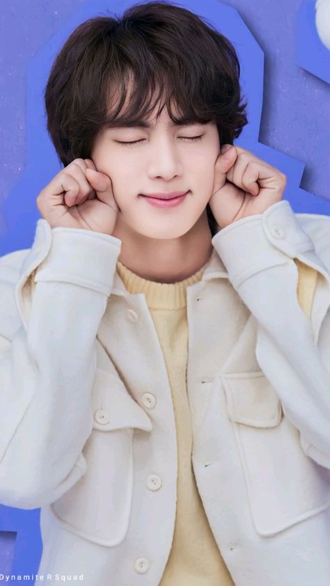 This contains an image of BTS Jin wearing a white jacket and posing with his hands on his ears