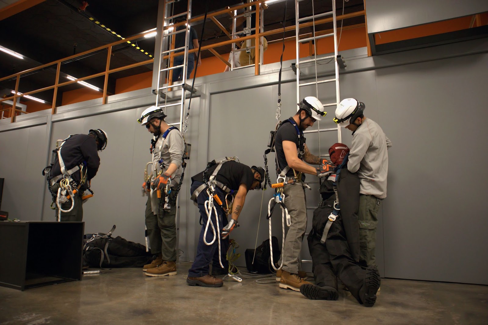 Five individuals in safety helmets and harnesses engaged in hands-on training, practicing rescue and safety techniques with equipment and mannequins. The industrial setting includes ladders and an orange railing, emphasizing a focus on technical skills and teamwork.